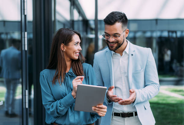 Shot of two confident colleagues walking and talking next to an office building. Businessman and businesswoman in meeting using digital tablet and discussing business strategy. Creative business persons discussing new project and sharing ideas while walking in front of their office building.